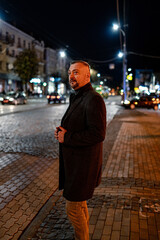 Obraz premium Mature fashionable adult man dressed in coat walking in urban setting. Stylish man walking on the street in the evening.