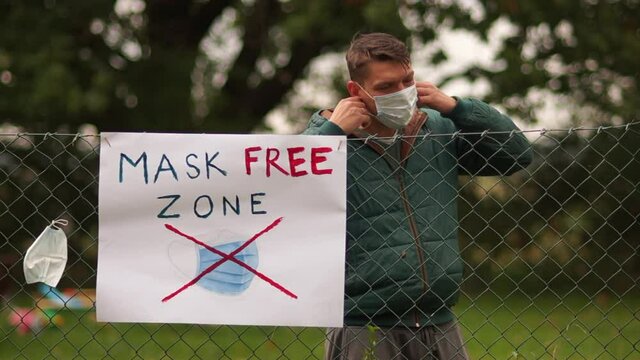 A poster on the fence with the inscription Mask Free Zone. A young male activist comes up and takes off his mask. I hung it on the fence next to the poster. Anti-mask concept, covid-denier