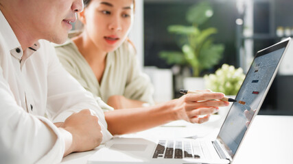 Asian business man present and explain work to female colleague, using laptop computer in office at night. Teamwork, coworker cooperation, financial marketing team, or corporate business concept.