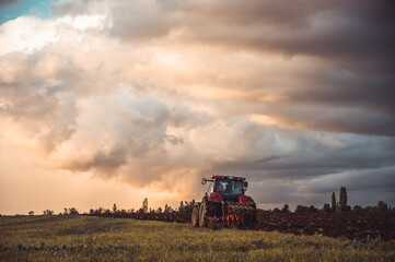 Fototapeta premium Tractor plowing the fields in the countryside by sunset. Agricultural tractor plowing the field. Red Tractor with plow. Sunset over the autumn field.