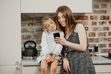 A European mother with pale skin is smiling while reading the news on a phone with a daughter in the kitchen. A mom is using a cellphone while her pretty girl is sitting on a countertop.