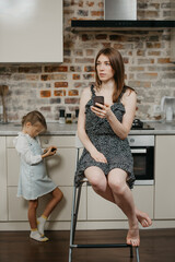 A mother with pale skin is holding a smartphone while sitting on a chair near a daughter which is gaming on a phone in the kitchen. A mom and her young girl are using their cellphones at home.