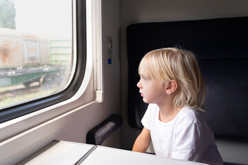 Fototapeta na wymiar Little blond boy looks thoughtfully out the train window. Rail travel with children