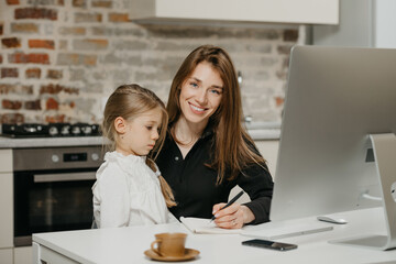 A young smiling mother is teaching a daughter at her remote workplace at home. A pretty mom is describing to a blonde child some things at an apartment.