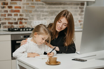 A young mom is hugging a pretty daughter while she is drawing in the notebook at home. A gorgeous mother is looking at her blonde child which is doing homework.