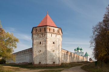 Fototapeta na wymiar A whitewashed tower with visible red bricks and a wall around a Russian monastery. White church with green domes in the background. Yellow and red trees around. Blue sky. No people.