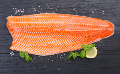 Half raw salmon fillet, salt and basil on black wooden table