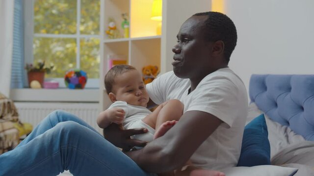 Cute newborn baby lying in arms of african father sitting on bed