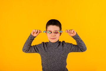 Jolly toddler boy covered his eyes with candy on a stick on a yellow background with space for text