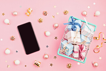 Christmas gift boxes in shopping basket and cell phone on pink background. Winter holidays online gift shopping