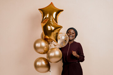 Christmas celebration. African american holiday. Attractive girl with golden helium balloons isolated peach background. Young black woman weared traditional dress and scarf.
