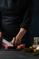 The chef in black uniform cuts with knife raw beef or pork on chopped wooden board at restaurant kitchen on dark blue background. Backstage of cooking dinner with meat, vegetables and spices. Closeup.