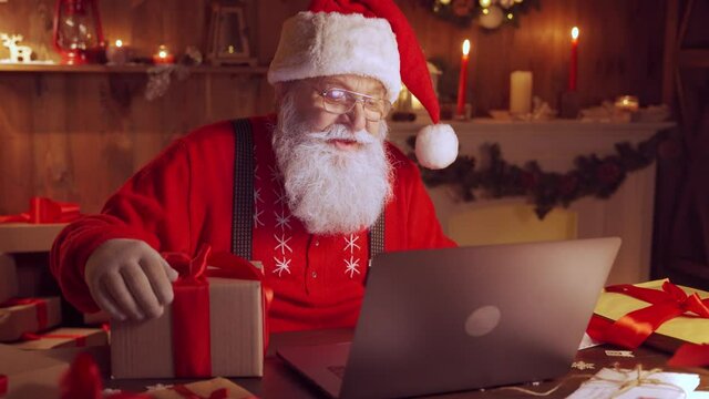 Old Santa Claus holding gift box video calling kid talking to child in virtual video online chat meeting on laptop sit at home table late saying greetings on xmas eve. Merry Christmas social distance.