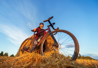 Naklejka premium Cyclist stands by a haystack in a field holding his bike and enjoying the magnificent sunset. Active lifestyle concept. Low angle view.