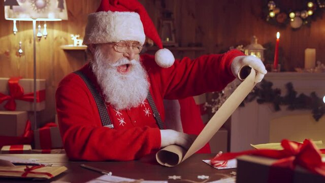 Happy elderly Santa Claus wearing hat holding parchment roll reading letter wish list sitting at home table late with presents and candles preparing for holiday on xmas eve. Merry Christmas wishlist.