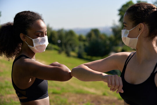 Two Diverse African And Indian Fit Ladies Friends Wear Face Masks Give Elbow Bump. Sporty Young Women Non-contact Greeting Together Outdoor Meeting For Fitness Training Or Running Outside. Closeup.