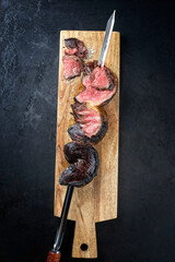 Barbecue dry aged wagyu Brazilian picanha from the sirloin cap of rump beef sliced and offered as top view on a skewer on a wooden design board on rustic black background with copy space