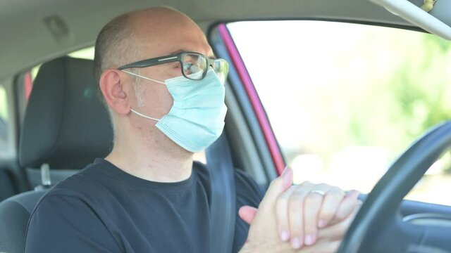 A middle-aged Caucasian man is in the car,  he wears the mask to protect himself from the coronavirus, sanitizes his hands by spraying disinfectant on the palm and then rubbing them together.