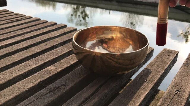 A man practices playing a Tibetan healing bowl on a meditation boardwalk near a pond