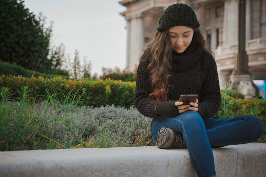 Adolescente sentada en las calles de la Ciudad de México en una tarde de otoño jugando con su teléfono celular, chica guapa posando sentada con ropa de abrigo,  covid19