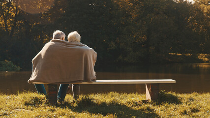 Elderly gray haired couple siting on the bench near the river covering with blanket on autumn day....