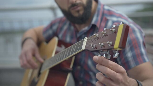 Close-up of guitar fingerboard with blurred bearded man playing musical instrument. Unrecognizable Caucasian musician guitarist performing on cloudy summer day sitting on urban bench.