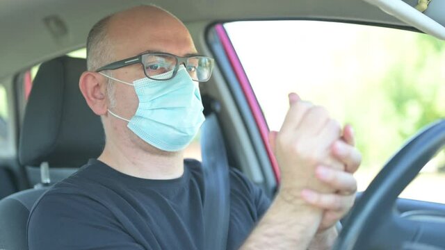 A middle-aged Caucasian man is in the car,  he wears the mask to protect himself from the coronavirus, sanitizes his hands by spraying disinfectant on the palm and then rubbing them together.