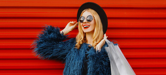 Portrait close up of stylish blonde woman with two shopping bags wearing a blue faux fur coat, black round hat over red wall background