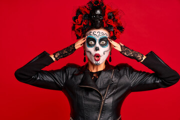 Girl with sugar skull makeup with a wreath of flowers on her head and skull, wearth lace gloves she keeps hands near her head and open shocked mouth isolated on red background. concept of Halloween