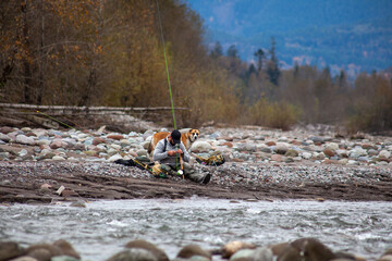 A man sits on the side of the Squamish River with his fishing equipment, changing a fly, with his dog trying to catch a fish in British-Columbia
