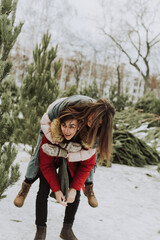 A young couple is fooling around, having fun in winter against the background of the Christmas tree market in the city. A young woman on the back of her boyfriend laughs and smiles