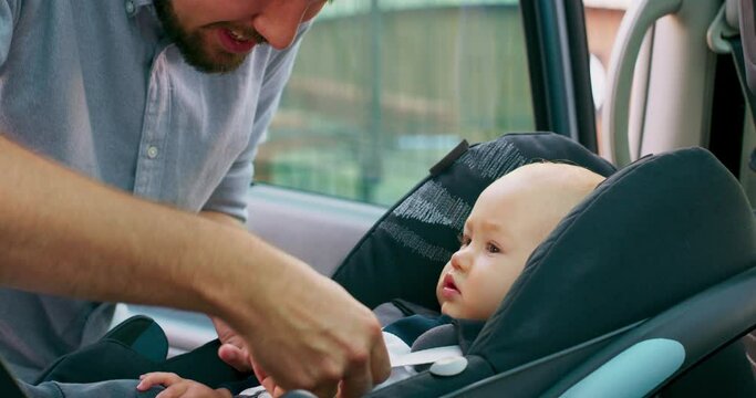 Camera inside the car. Closeup baby boy sits in the baby car seat inside of car. Young bearded father checks baby's fastened seat belts, and talks to him. Slow motion