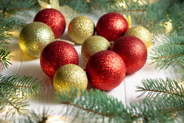 Green branches of a Christmas tree with light bulbs and shiny golden and red balls on a white wooden background.