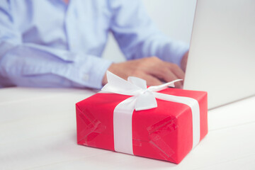 Hand of man holding red gift box and having laptop computer on wooden table, online shopping for giving present for Christmas day or anniversary, season and celebrate during social distancing.
