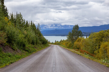 View of Scenic Road alongside Lake, surrounded by Trees and Mountains on a Cloudy Fall Day in Canadian Nature. Taken in Northern British Columbia, Canada.