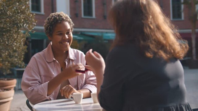 Woman holding engaged ring and making proposal to girlfriend having date in outdoors cafe