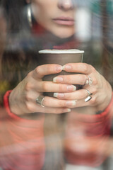 close-up of a person's face holding a cup of coffee with his two hands, in his fingers he has several rings, photo behind glass
