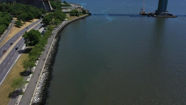 An aerial shot of the shores of Lower New York Bay near Shore Parkway in Brooklyn. It is a sunny day as the camera dolly in, looking down over a path & the water. A bridge tower's base comes into view