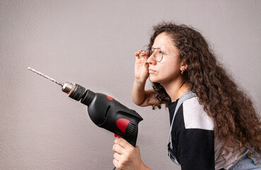 Mujer joven trabajadora sosteniendo un taladro mientras se arregla los lentes.  Taller de...