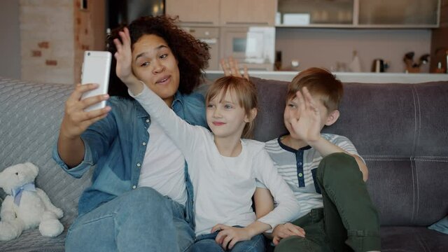 Joyful kids boy and girl are making online video call waving hand sitting on couch with nanny holding smartphone enjoying modern communication