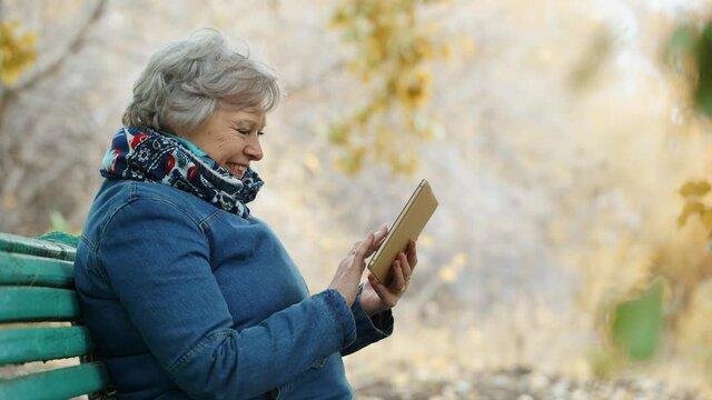 Elderly woman walks in the park in autumn using a digital tablet, online in social networks.