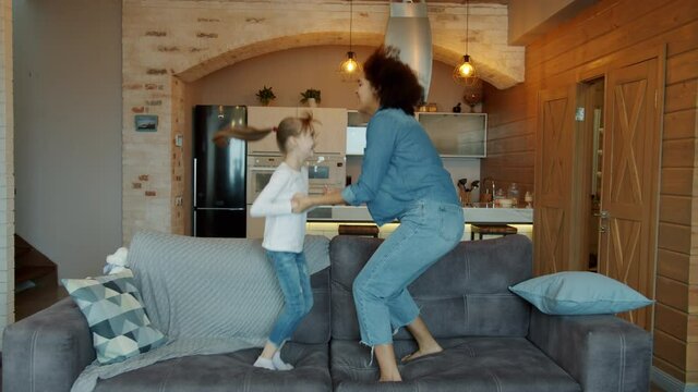 Playful girl and Afro-American woman babysitter are jumping on sofa in cozy room dancing having fun holding hands. Childcare and babysitting concept.