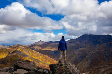 Fototapeta na wymiar Man Standing and Looking at Viewpoint of Mountain Range on a Cloudy Fall Day in Canadian Nature. Taken in Tombstone Territorial Park, Yukon, Canada.