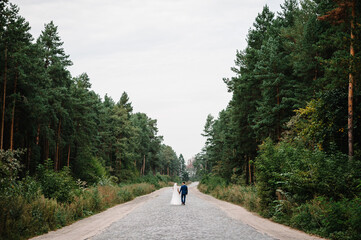 Wedding couple walking in nature. The bride with wedding bouquet and groom go on the pavement road and hold hands on a stroll along forest. Newlyweds of the outdoors. Back view.