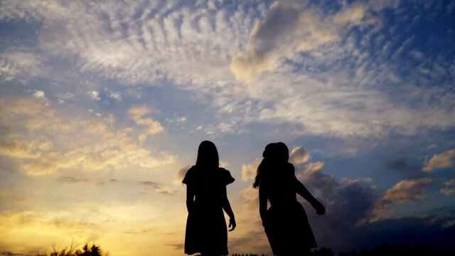 Two young woman with long hair , in dresses, jumping and having fun , on a summer evening. Their silhouettes are visible against the blue sky with white clouds. Sunset, the last rays of the sun.