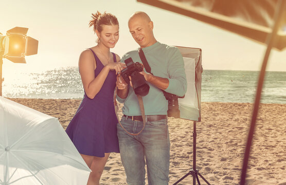 Professional adult photographer showing photos on digital camera to smiling model girl during shooting on sea coast