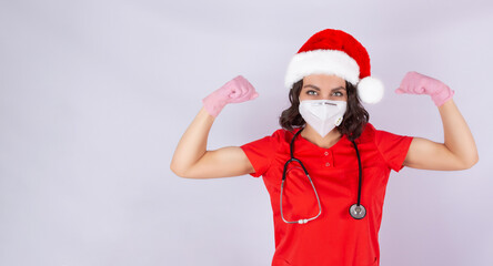 A happy doctor woman in a Santa hat and protective mask shows a winning gesture, strength.  Isolated on a white background. Girl medical worker in a new year's hat.