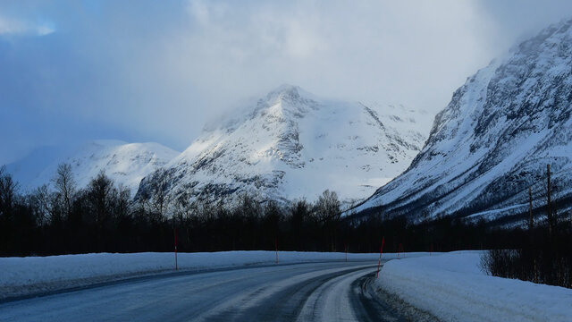 Cleared rural road with snowplow markings (red posts), bare trees and snow-covered mountains in background between Fagernes and Brevikeidet, Troms og Finnmark, Norway with cloudy sky in winter time.