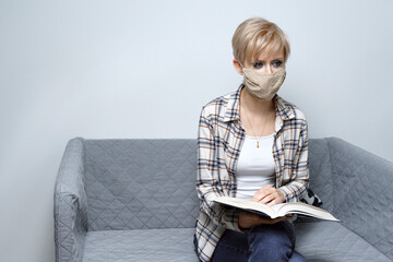 blonde woman in protective mask reading a book sitting on the couch. obtaining a second education...