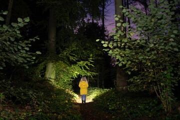 Girl with a yellow coat standing in the dark forrest, searching the path with a flashlight